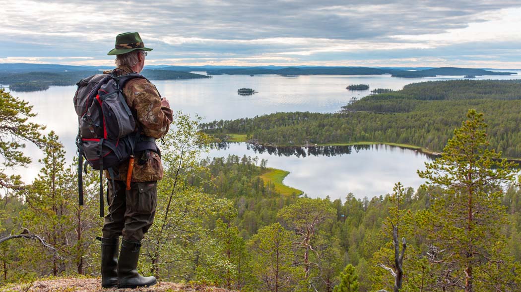 The hiker with backpack standing at a viewing point on top of a tall hill. A lush and summery lake- and forest landscape can be seen from the hill. The sky is cloudy.