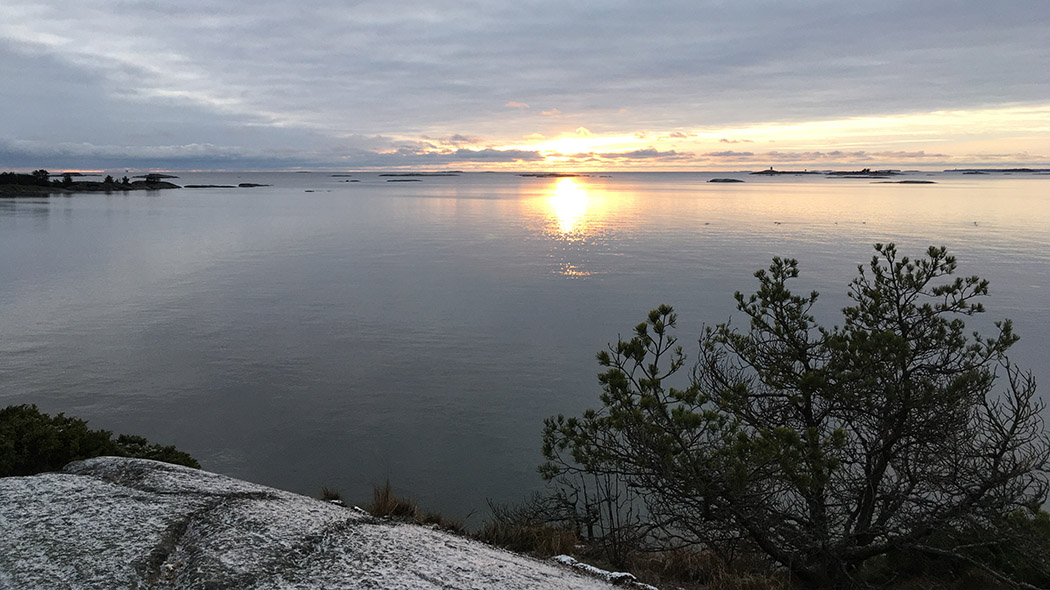 Seaside, sea and islands further away. In a mostly cloudy sky, the sun shines low.