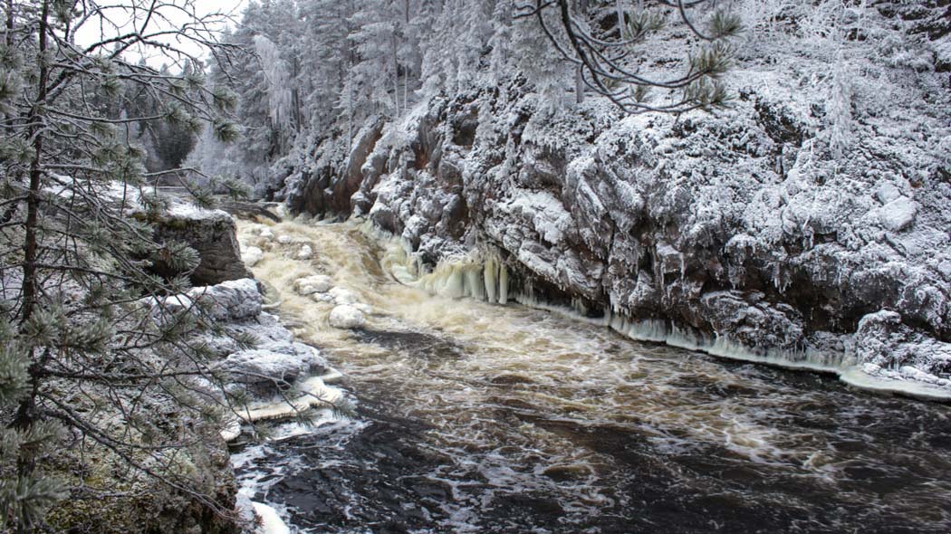 Red rock cliffs topped with a thin layer of snow. Between the cliffs, a frost-covered rapids roars.