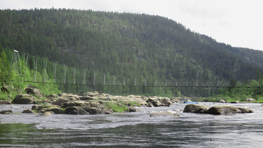 A fast-flowing river with rocky banks and rising land on both sides. A hanging bridge crosses the river.