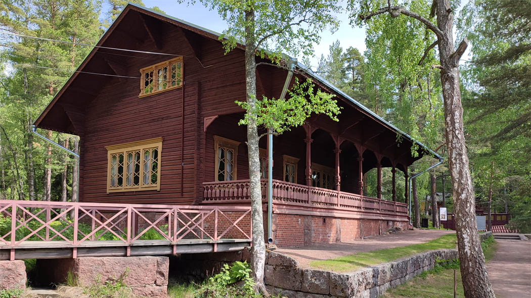 A dark wooden house on two floors with a large veranda in the foreground.