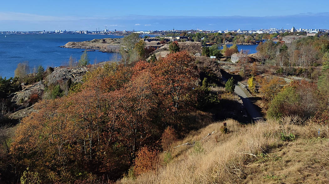 Autumn landscape from a high grassy slope, in the foreground sea, trees, shrubs and open rock, as well as a narrow road. In the background the city's tall houses, towers and chimneys.