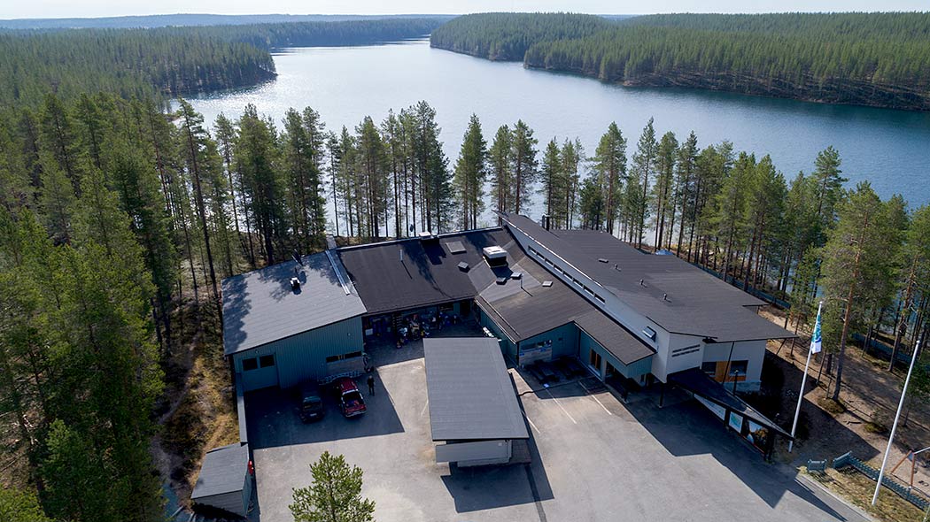 An aerial phtograph of a building, cars are parked on an asphalted yard. A forest surrounds the building.