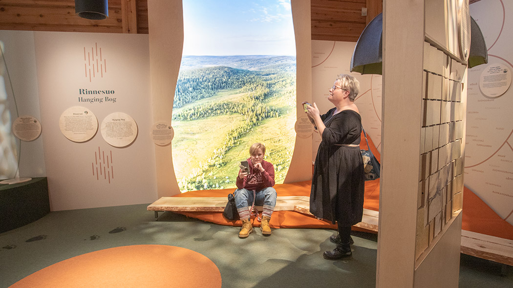 Two people are in the exhibition. One is sitting with a mobile phone in front of a large landscape picture and the other is taking a picture of the exhibition with his phone.