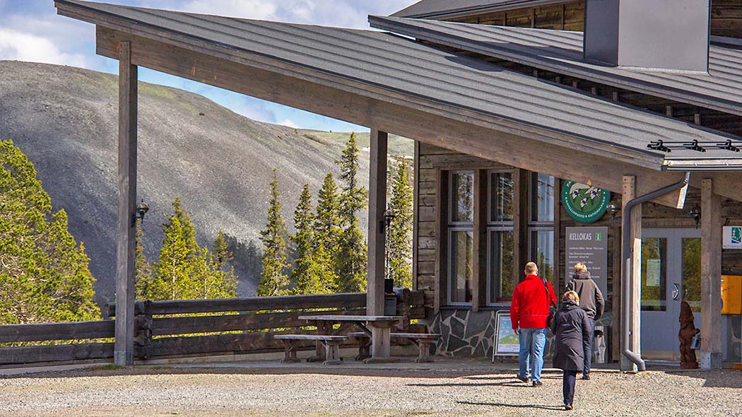 Three hikers walking towards a modern wooden building on a sunny day. There is a terrace in front of the building. A forest and a stony fell can be seen in the background.